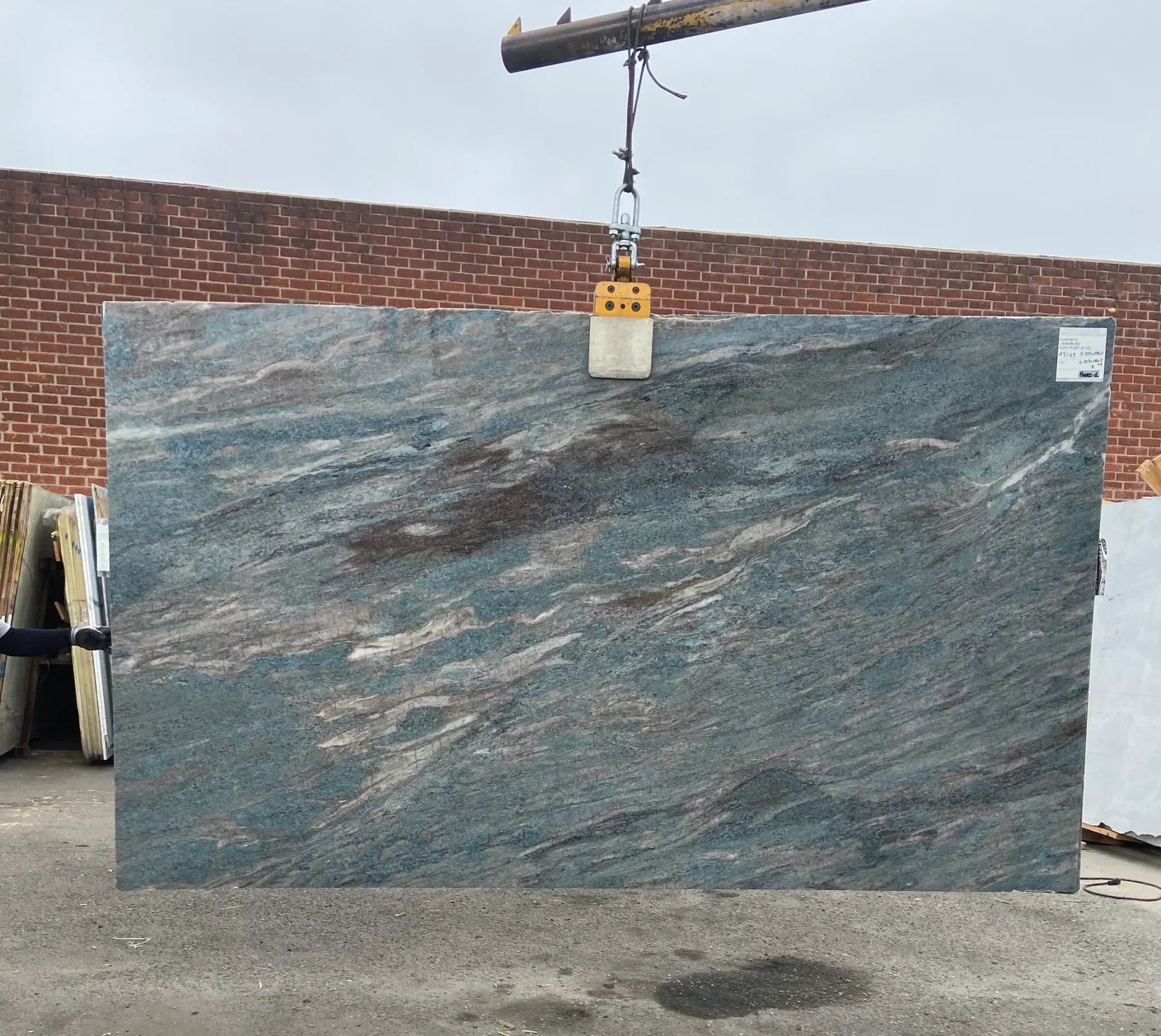 Antolini Caribbean Blue quartzite slab, 79 by 134 inches, polished finish with soft sea-blue field swept by diagonal currents of cream, rust, and burgundy, lifted by crane against a brick wall at Royal Stone in Los Angeles