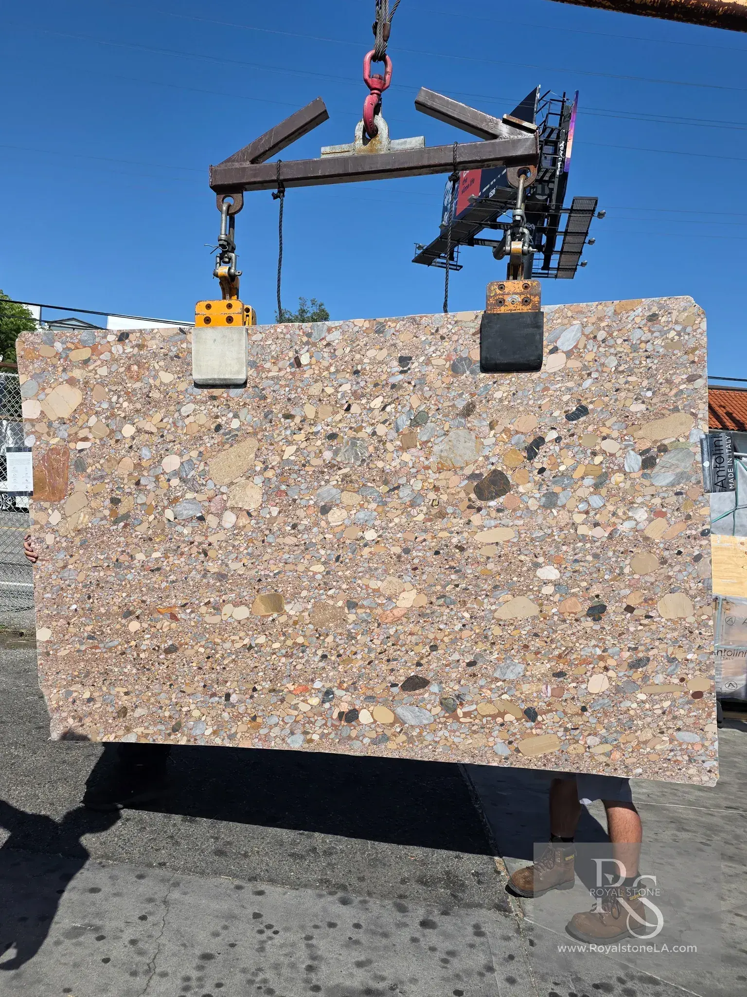 Antolini Ceppo Rosa di Palladio honed marble slab, 62 by 101 inches, dense pink-rose and cream field packed with rounded pebble fragments in grey, ochre and charcoal, lifted by crane under blue sky at Royal Stone in Los Angeles