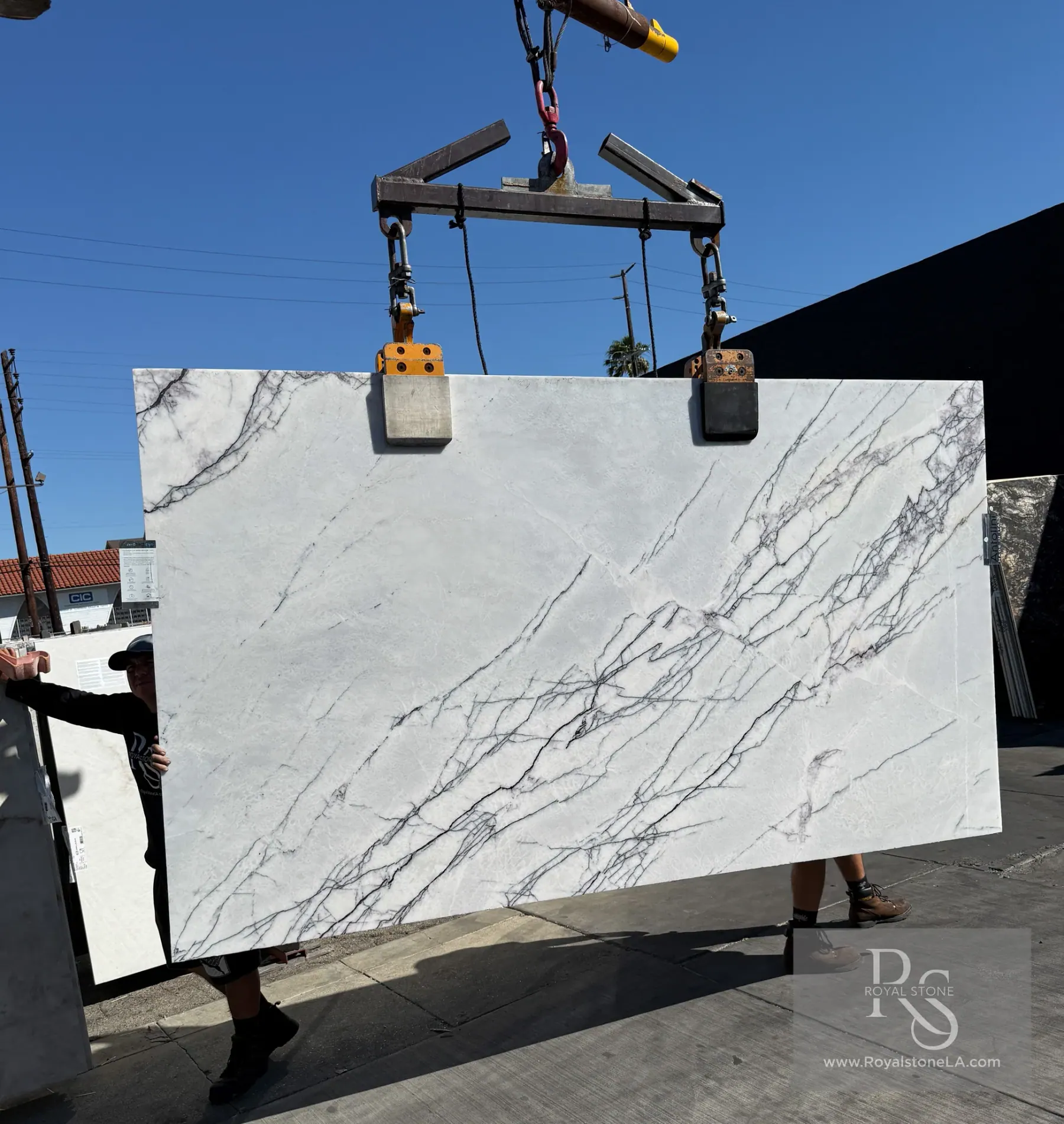 2cm Antolini Lilac marble slab, cut EE007, 69 by 121 inches, polished finish with bright white field crossed by a sharp diagonal network of violet, plum and near-black veining, lifted by crane under blue sky at Royal Stone in Los Angeles