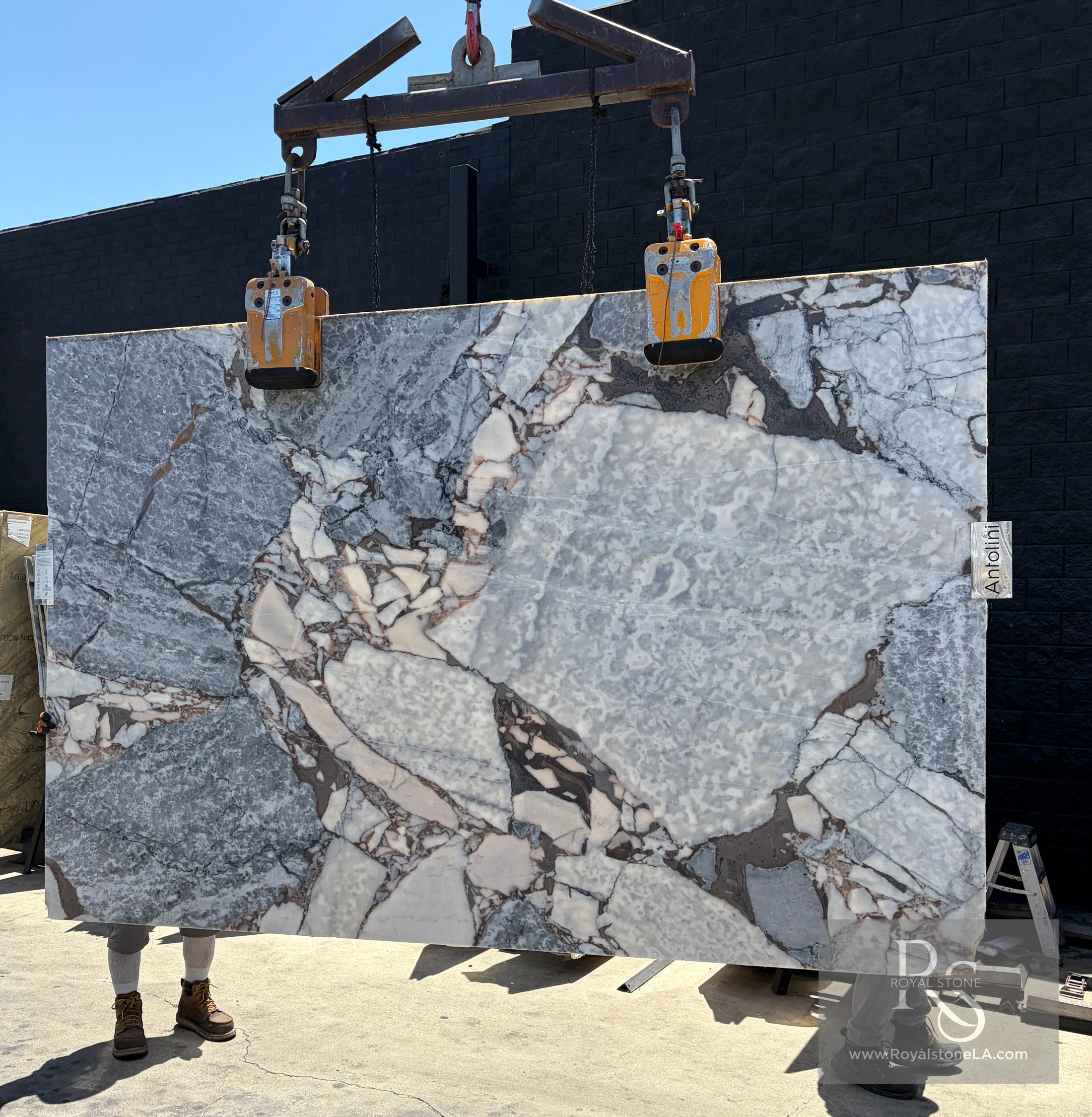 Antolini Ocean Storm Viola marble slab honed, lifted on overhead crane at Royal Stone Los Angeles — steely blue-grey ground shattered by white breccia clasts, rose and amber fragments, and a large central translucent quartz pocket, 73 by 111 inches