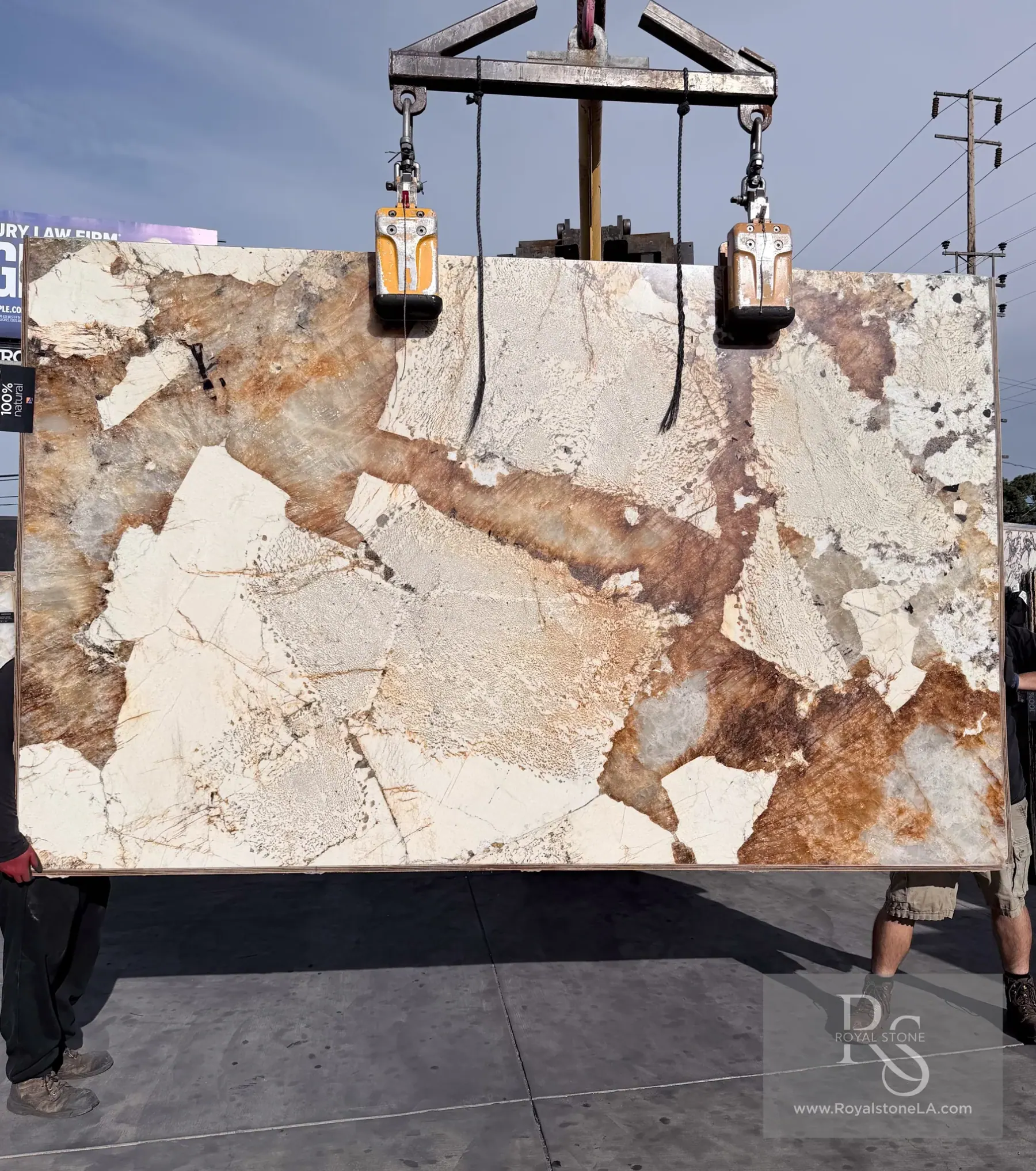 2cm Antolini Patagonia Original Unique AO683 polished quartzite slab, 75 by 121 inches, ivory and cream fragments scattered across a cool ground with amber, rust and burnt-orange veining running diagonally between them, lifted by crane against a partly cloudy sky at Royal Stone in Los Angeles