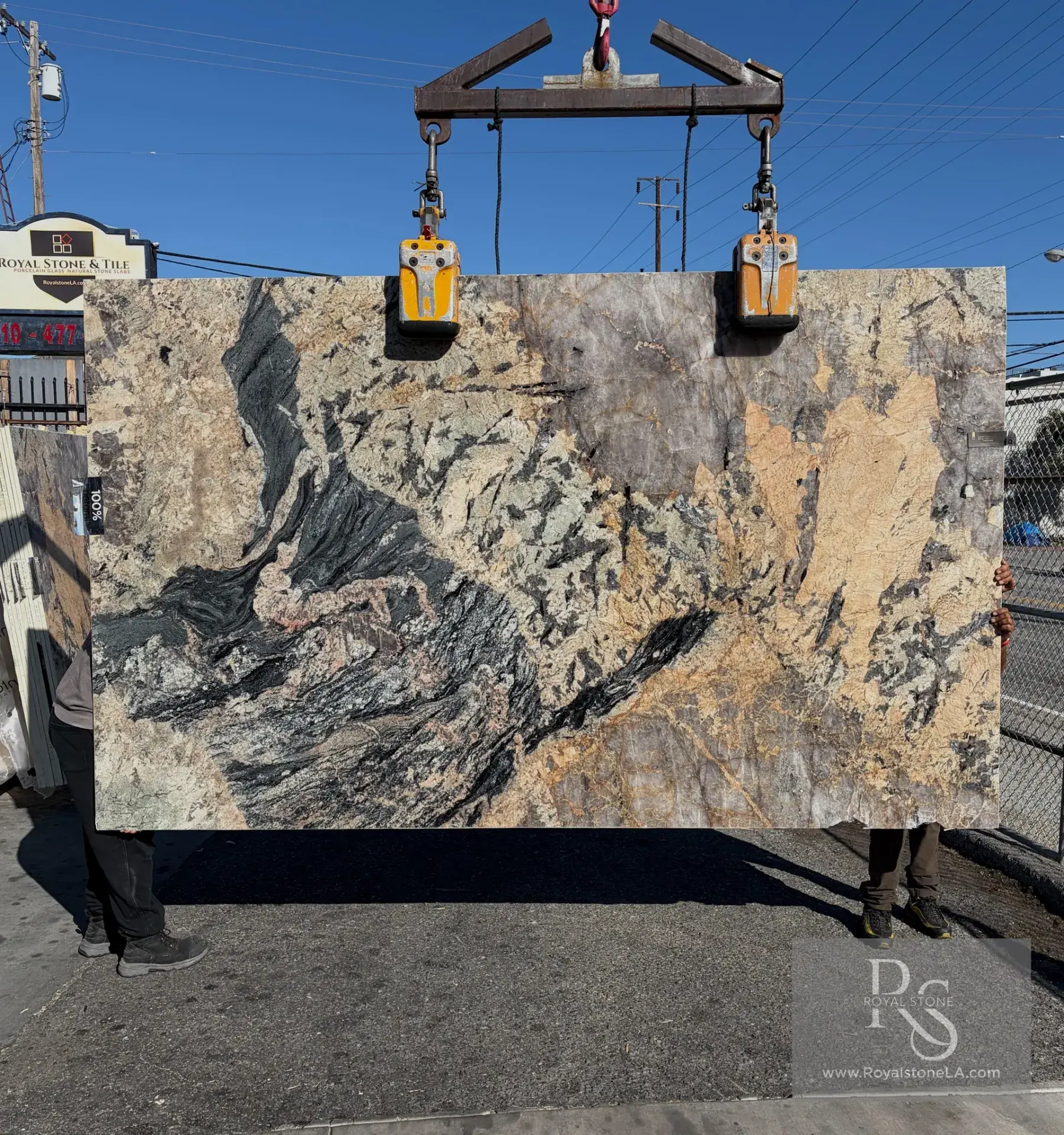 2cm Antolini Patagonia Tourmaline Unique IYA343 leathered quartzite slab, 75 by 125 inches, charcoal mineral streaks sweeping across a warm peach and gold crystalline ground with pockets of pale grey quartz, lifted by crane in front of the Royal Stone sign in Los Angeles