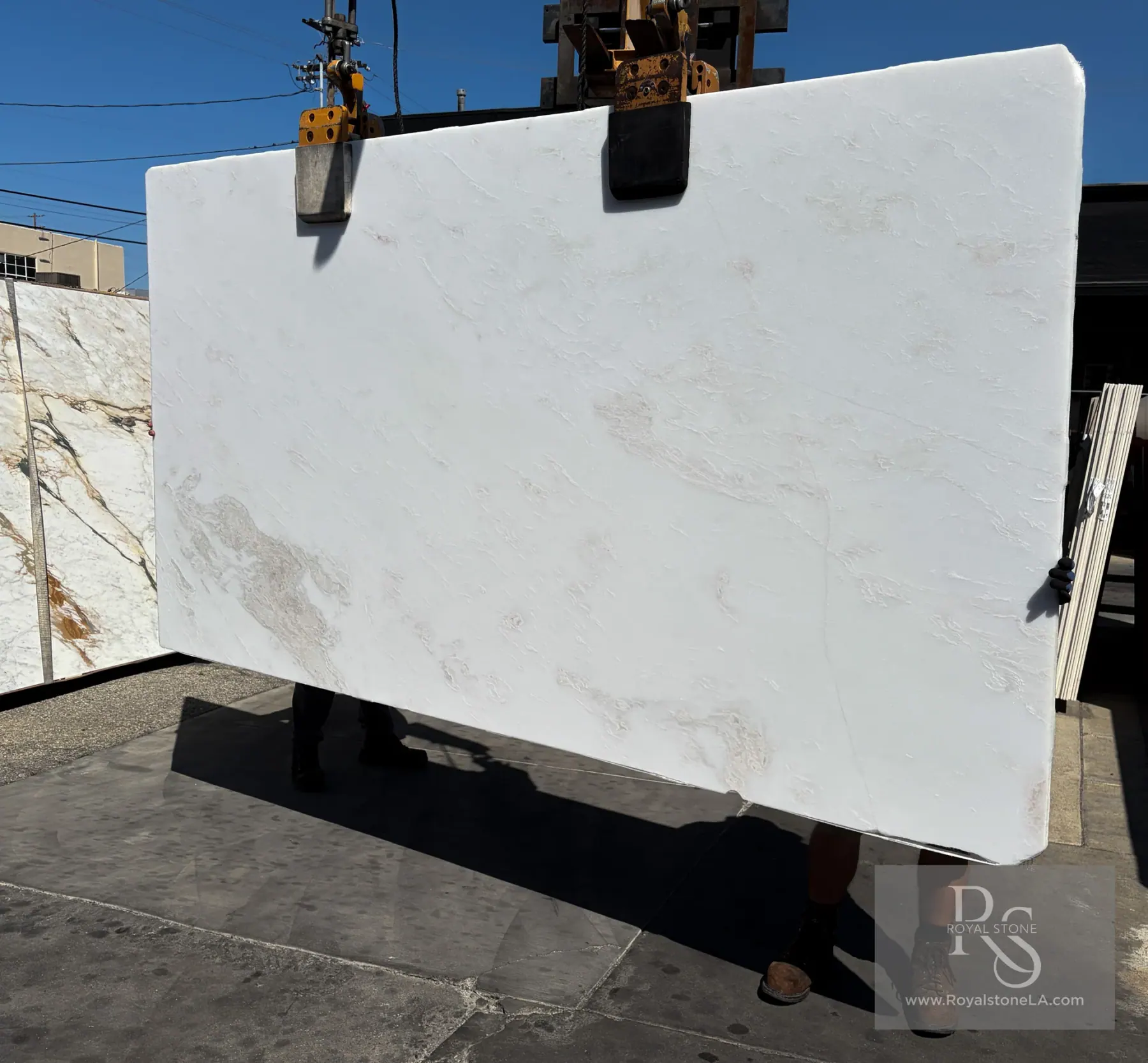 2cm Bianco Namibian Rhino White marble slab, 65 by 122 inches, honed finish, almost vein-free pure white field with the faintest grey and warm beige drift, lifted by crane against a clear blue sky at Royal Stone in Los Angeles