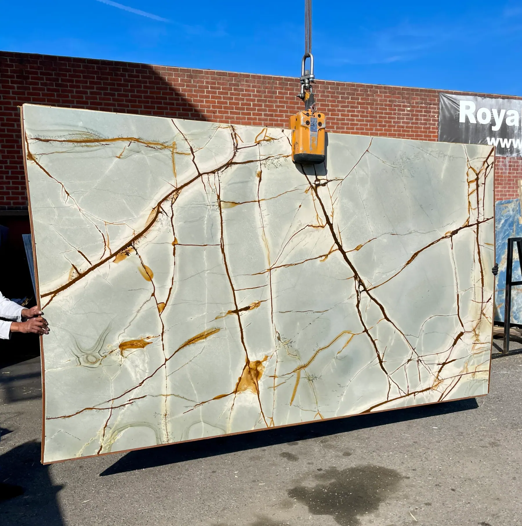 Blue Roma quartzite slab on the crane at Royal Stone, pale sage-blue field with rust-amber fracture network, polished finish