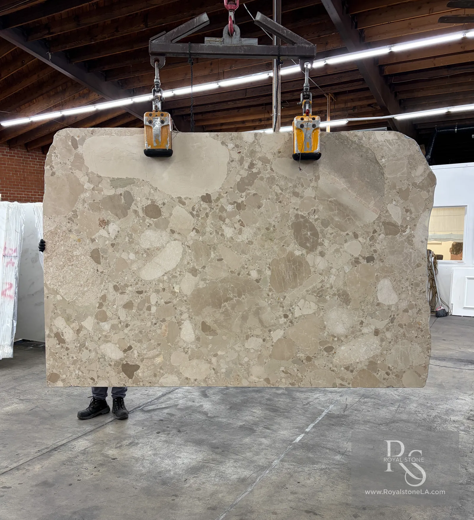 2cm Ceppo Creme marble slab, honed, 77 by 114 inches, soft cream ground crowded with large rounded oatmeal and taupe clasts, suspended from a crane clamp inside the Royal Stone yard in Los Angeles