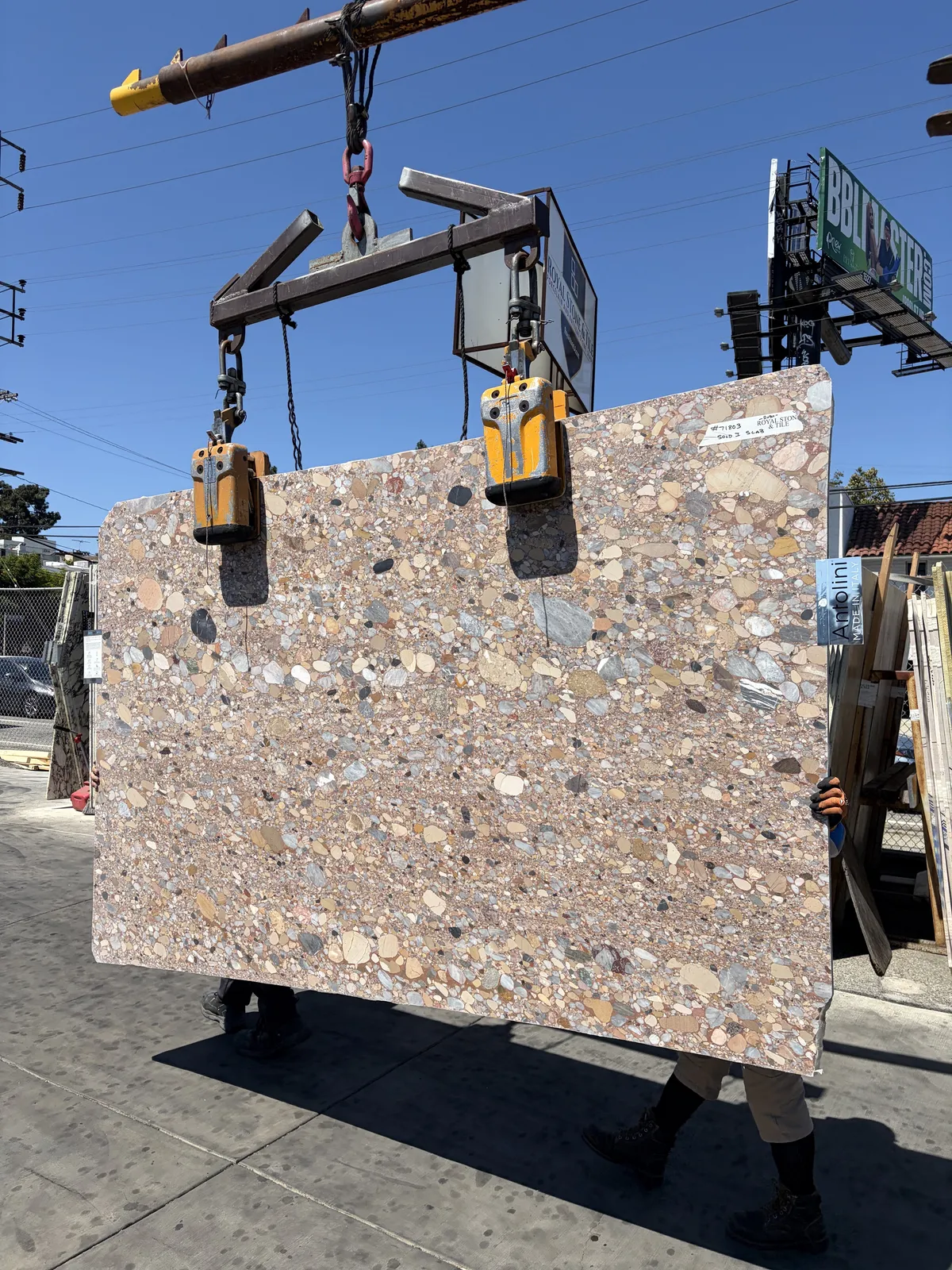 Full face of the Antolini Ceppo Rosa di Palladio honed slab, tag #71803, crane-lifted on a forklift boom in the Royal Stone yard, showing the dense conglomerate of rounded grey, ochre and cream pebbles across the warm rose ground