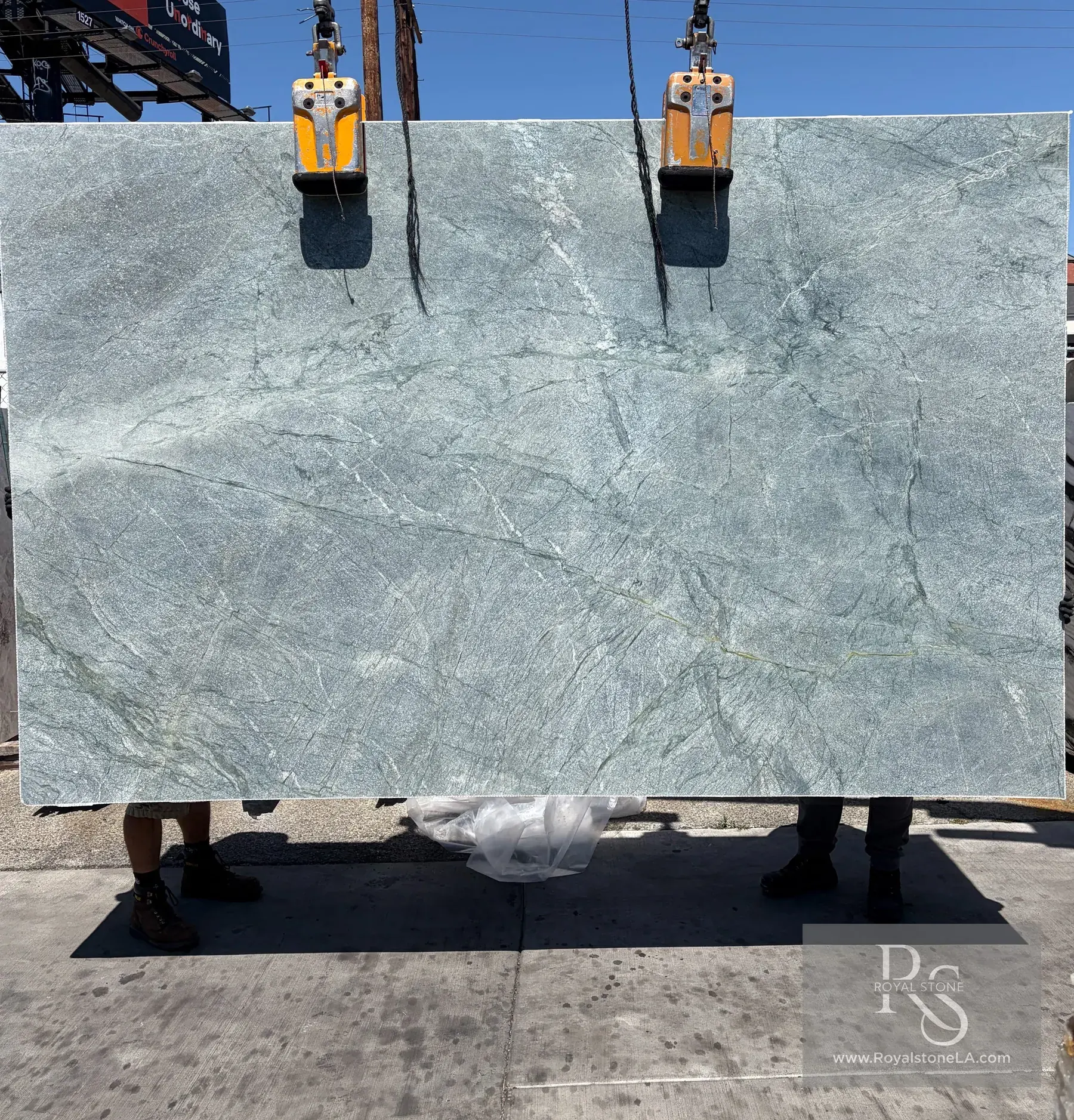 2cm Costa Smeralda quartzite slab, 79 by 123 inches, heavy leather finish, soft seafoam-green field with delicate white crystalline veining and a fine gold mineral trace, lifted on the vacuum gantry under blue sky at the Royal Stone Los Angeles yard