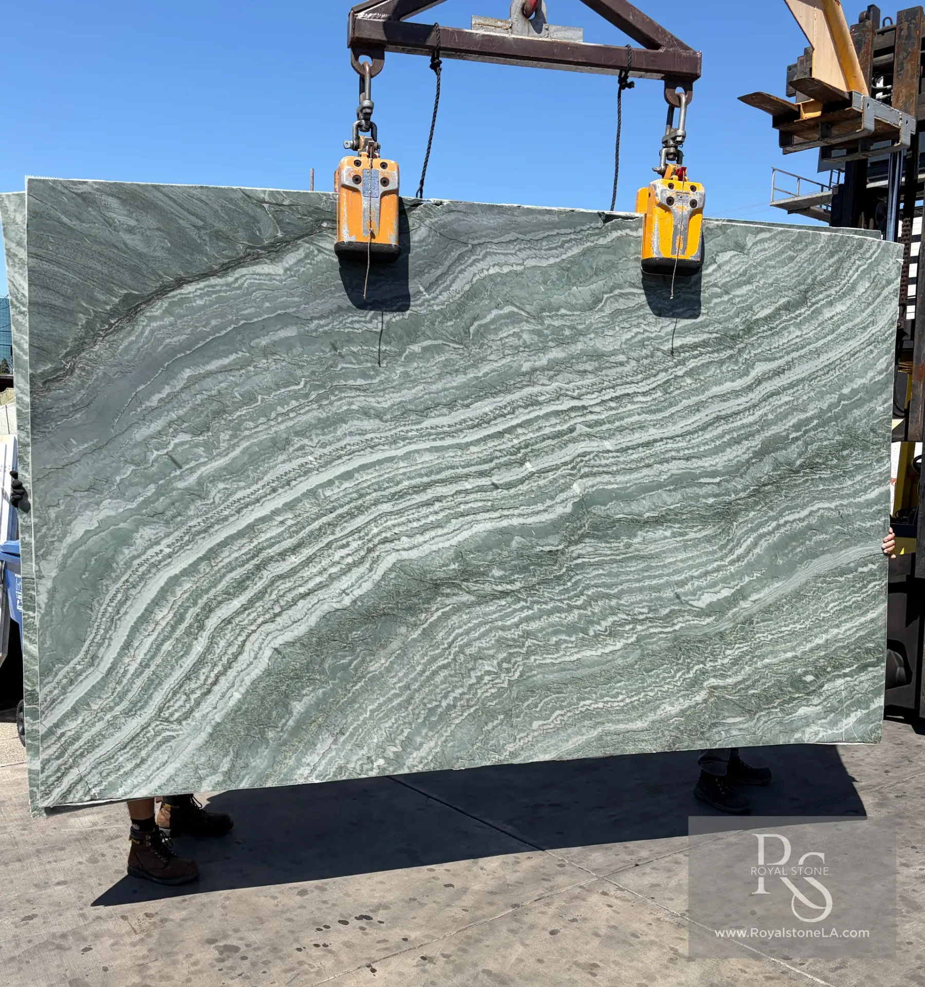 Emerald Quartzite slab, 76 by 121 inches, sage and jade field crossed by long sweeping horizontal striations of paler silver-green and ivory, lifted on the crane against a clear blue sky at Royal Stone in Los Angeles
