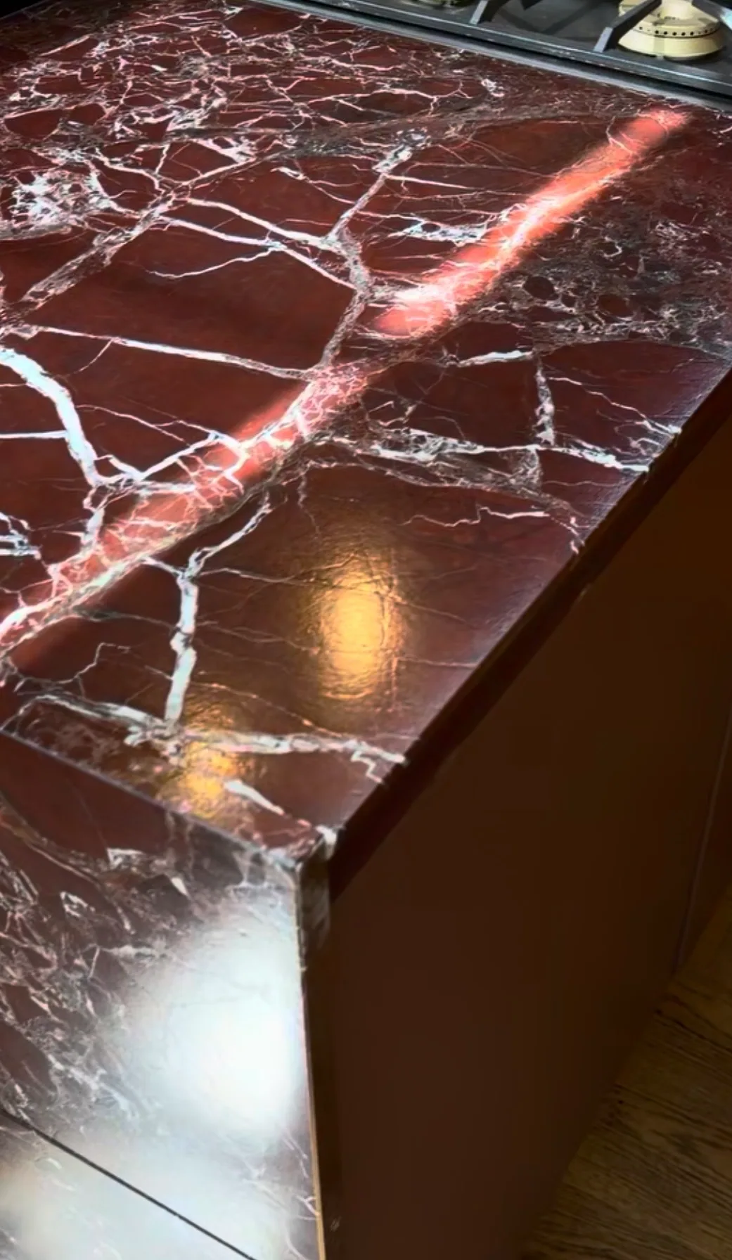 Close-up corner detail of Rosso Lepanto leathered marble countertop — deep oxblood field with bright white calcite veining continuing across a mitered edge, matte low-sheen surface with the veining sitting slightly proud of the red ground