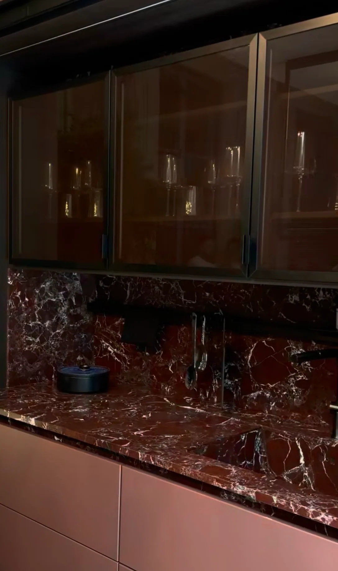 Rosso Lepanto leathered marble installed in a contemporary kitchen — countertop, mitered waterfall edge, and full-height backsplash all in continuous burgundy marble with white calcite veining, set against soft-pink lower cabinetry and bronze-framed reeded-glass upper cabinets, in low evening light