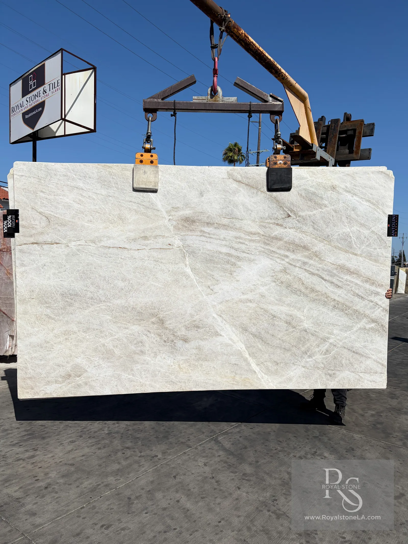 Taj Mahal Select leather-finish quartzite slab from Antolini block AO887 at Royal Stone Los Angeles, pale ivory field with soft horizontal drift, 78 by 131 inches, lifted by overhead clamps on the yard floor