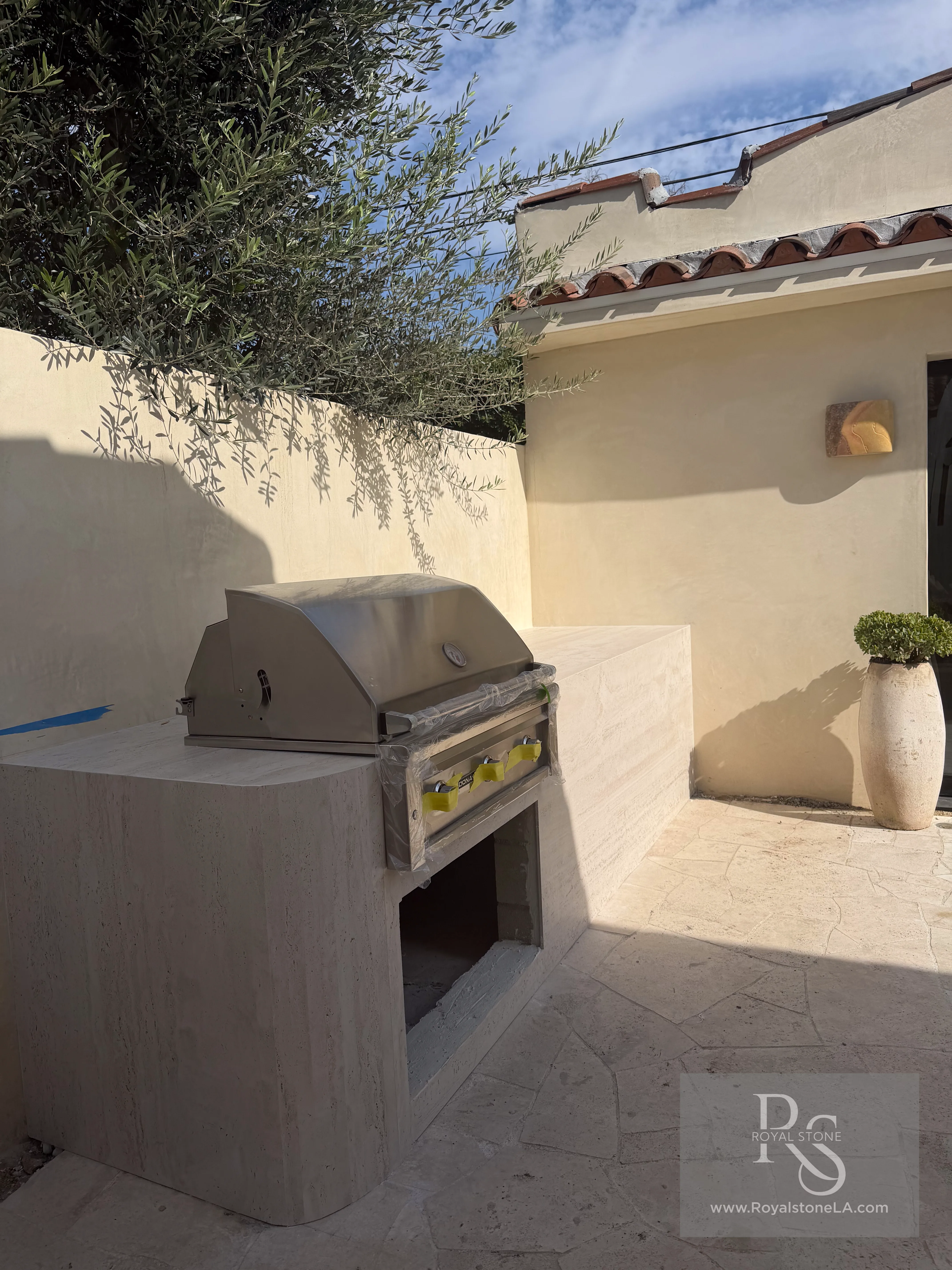 Installed outdoor BBQ island clad in ABK Navona Bone vein-cut 3D honed porcelain with a fully rounded curved end and waterfall to the floor, photographed in evening light