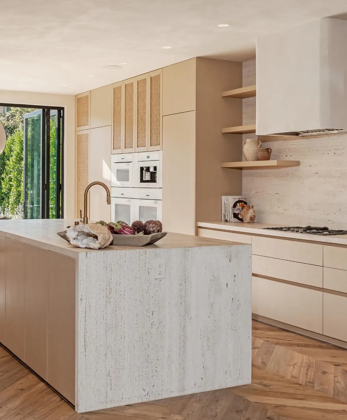 Minimalist kitchen with a monolithic Bianco Navona travertine waterfall island in an unfilled, sandblasted finish — soft ivory stone with open natural pitting — set against pale oak slab cabinetry, cane-front uppers, a plaster hood, and herringbone wood floors.