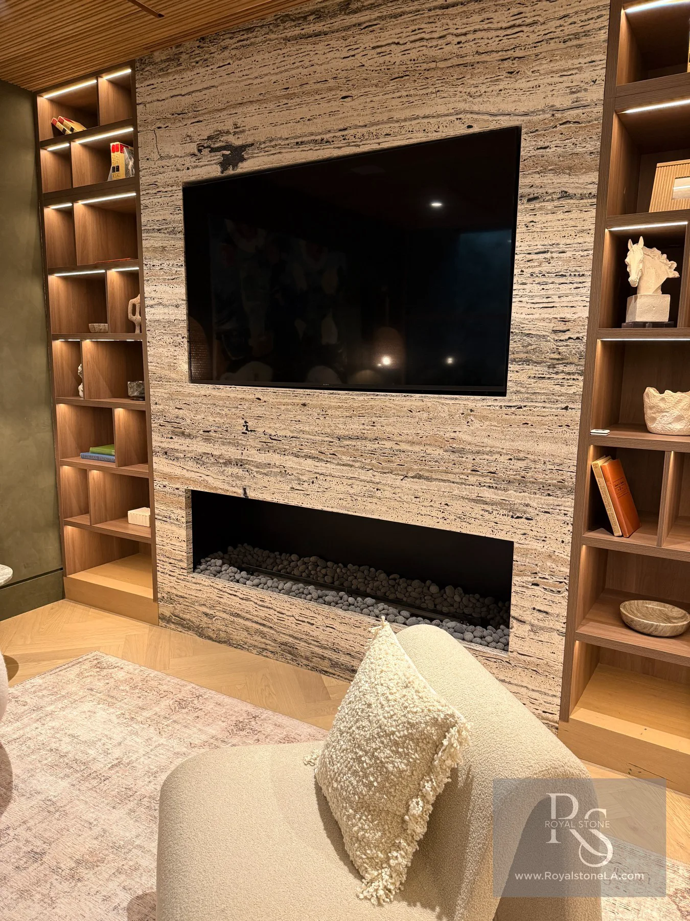 Living room media wall clad in Antolini Black Label Platinum Travertine, vein cut and leathered — a horizontal silver-grey and warm bone field with charcoal seams framing a wall-mounted Sony display above a long linear gas hearth filled with grey lava stones, flanked by warm walnut library millwork with concealed lighting, over a herringbone oak floor with a bouclé chair in the foreground.