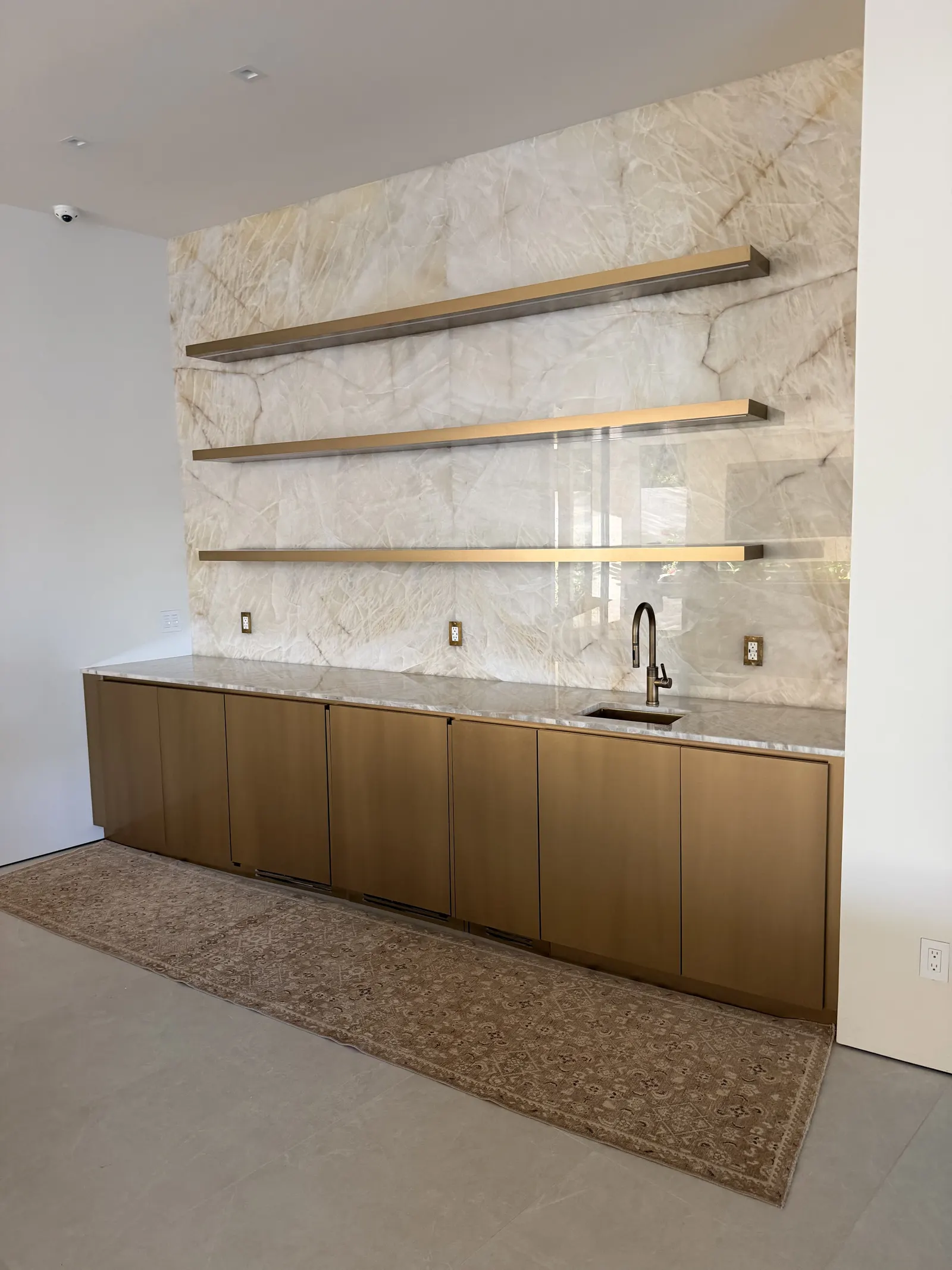 Full-height Cristallo quartzite wet bar wall in close detail — translucent crystalline white field threaded with amber and honey veins, three slim brass floating shelves cantilevered off the slab, an antique brass faucet, and oak millwork cabinetry below.
