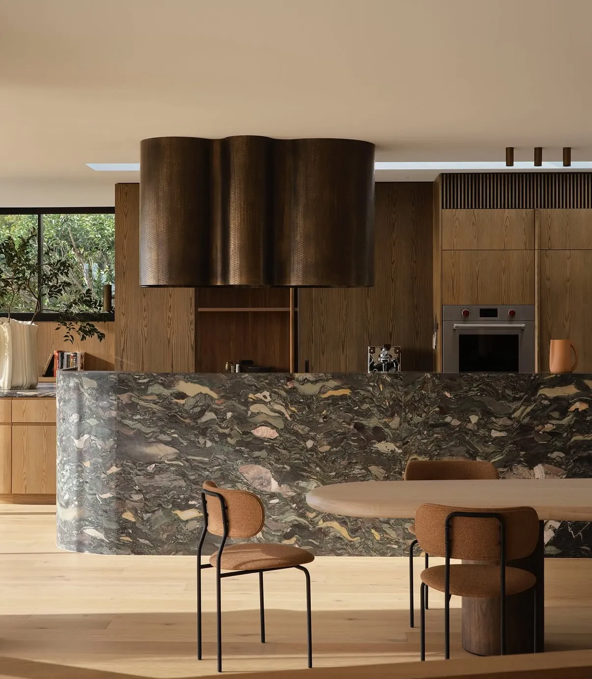 Curved waterfall kitchen island wrapped in Four Seasons marble — a dramatic multicolor field of olive green, plum, rust, and cream clasts flowing through dark grey stone — set against quarter-sawn walnut cabinetry, a hammered-bronze cloud-form hood, and an oak dining table with bouclé chairs.