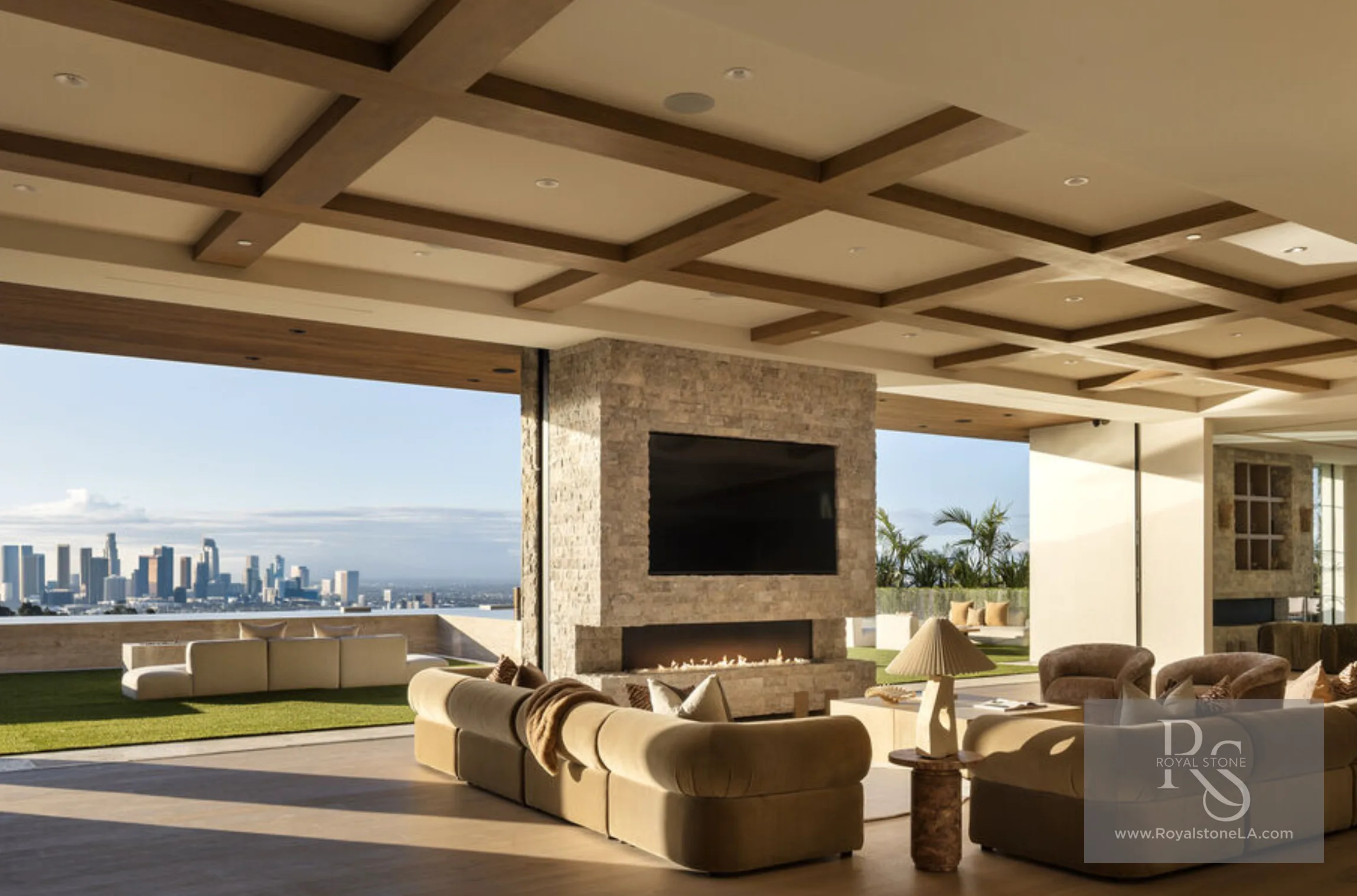 Indoor-outdoor loggia at 1360 Laurel Way in Beverly Hills — a freestanding cleft silver travertine column clad on all faces, carrying a recessed television above a linear fire, under a wood-beamed coffered ceiling, with pocketing glass walls fully open to a lawn and the downtown Los Angeles skyline beyond.