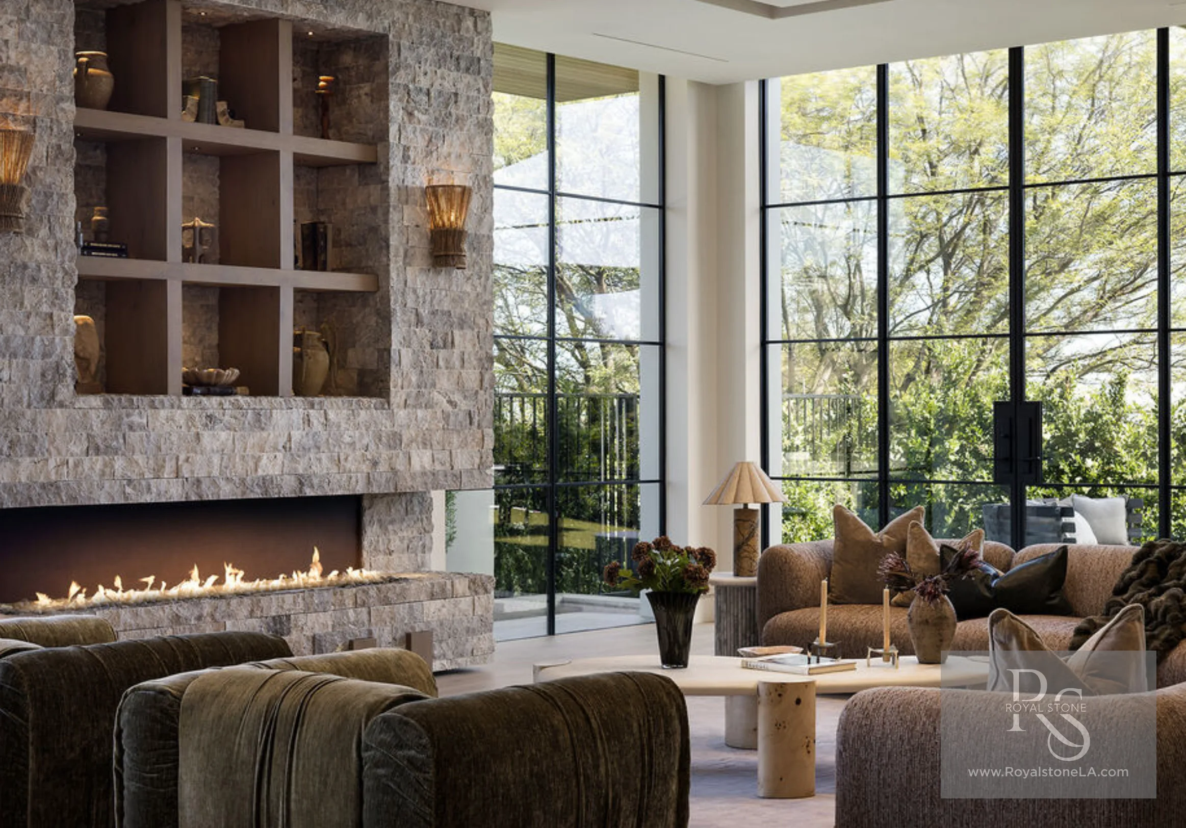Double-height great room at 1360 Laurel Way in Beverly Hills — a full-height split-face silver travertine fireplace with a long linear fire and a recessed grid of niche shelving holding earthenware vessels, set against floor-to-ceiling steel-and-glass window walls that look onto mature trees, with chenille and bouclé seating arranged below.