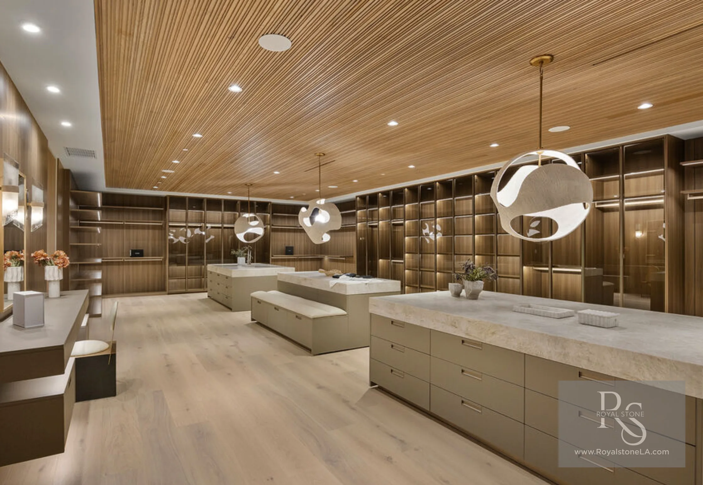Expansive dressing room at 1360 Laurel Way in Beverly Hills — three monolithic islands topped in Taj Mahal quartzite with thick mitred edges, surrounded by floor-to-ceiling walnut wardrobe cabinetry, a slatted wood ceiling, and sculptural pendant lights.