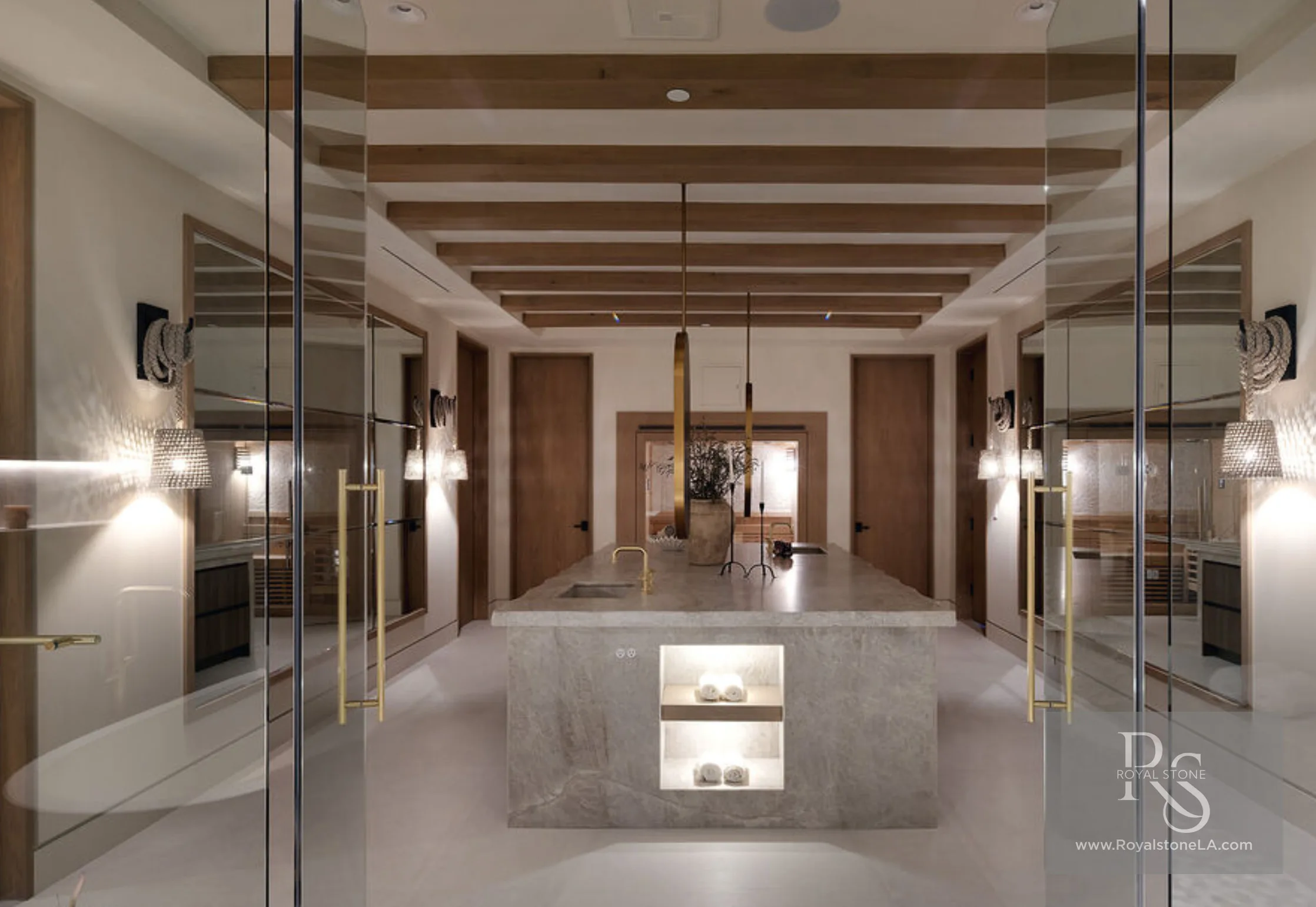 Symmetrical spa bath at 1360 Laurel Way in Beverly Hills — a central monolithic Taj Mahal quartzite island with a lit display niche of rolled towels and brass fittings, flanked by mirrored vanity walls and wood-beamed ceilings, framed through frameless glass partitions.