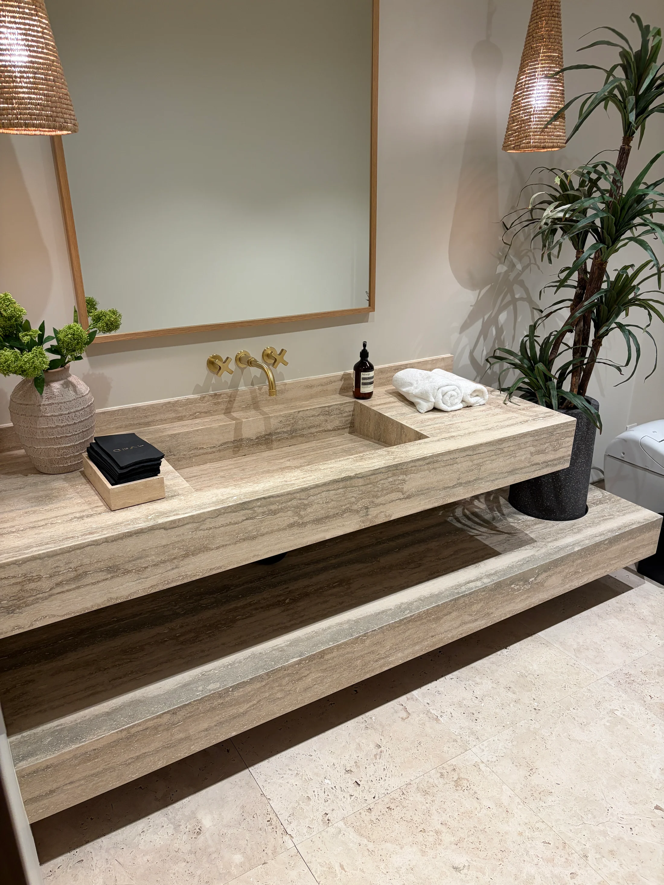 Foyer at 1360 Laurel Way in Beverly Hills — a long cream travertine console carved as one block, holding a small olive-tree planting, an aged-brass vase, and a wax-sculpture vessel, set on a pale oak chevron floor against a softly tinted plaster wall and an antique mirror.