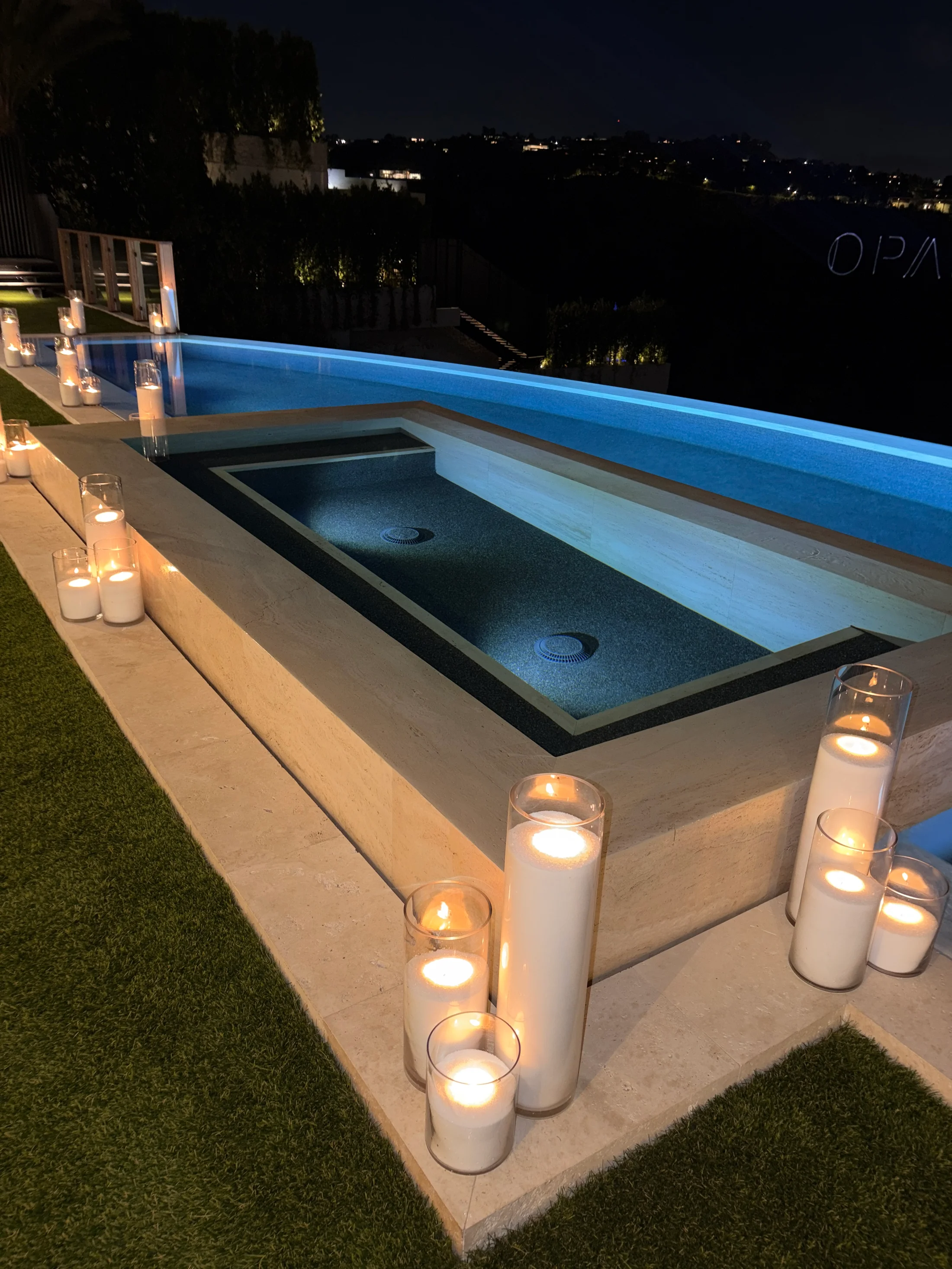 Outdoor terrace at 1360 Laurel Way in Beverly Hills at dusk — a long travertine deck lined with clustered glass-cylindled candles leading toward a recessed fire feature, with the downtown Los Angeles skyline and lit treetops visible beyond the lawn.