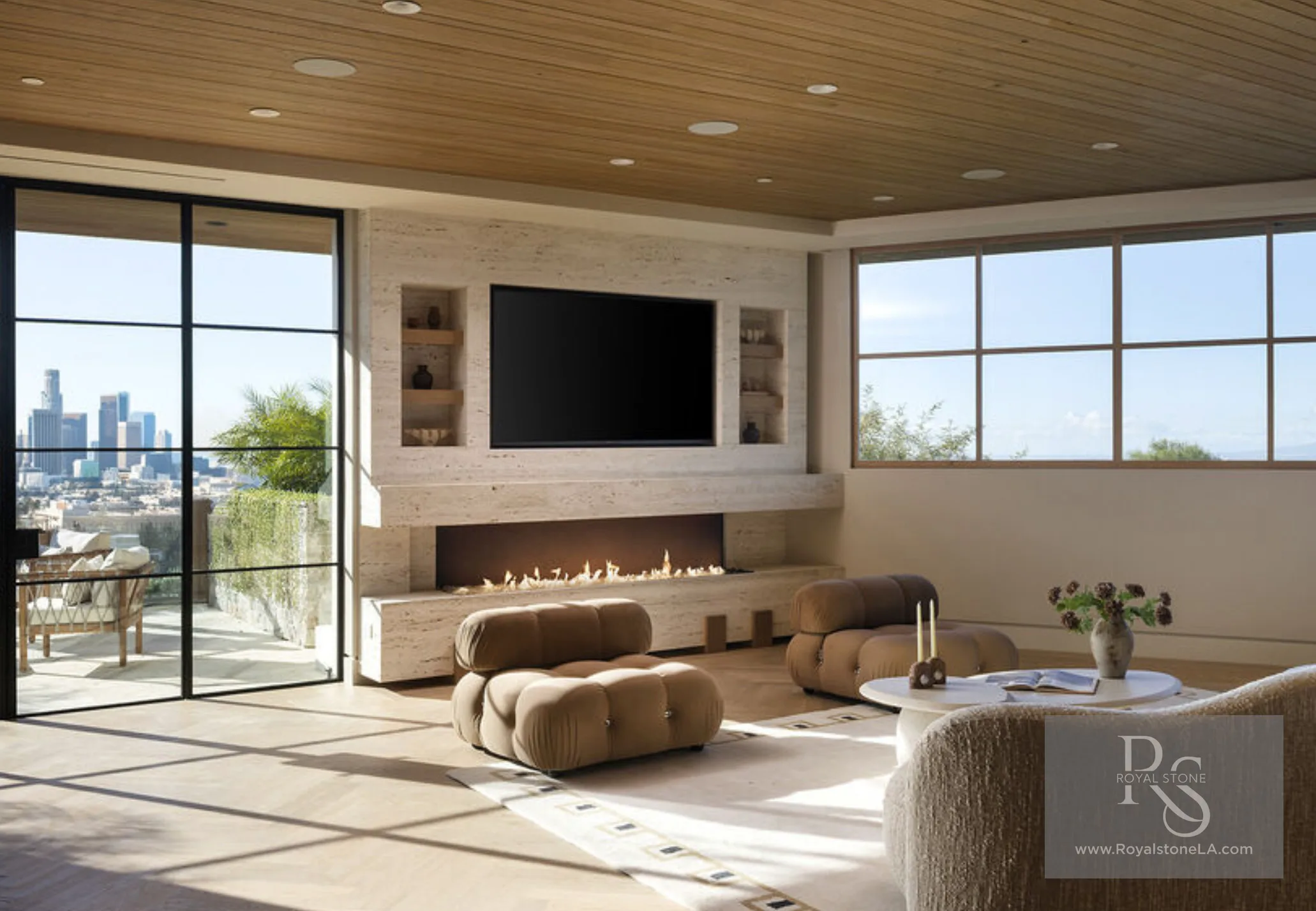 Sunlit lounge at 1360 Laurel Way in Beverly Hills — a vein-cut travertine media wall with a wall-mounted television above a long linear fire and a floating hearth bench, flanked by inset niche shelving, with a slatted wood ceiling, modular tan seating, and steel-framed glass walls opening to the downtown Los Angeles skyline.