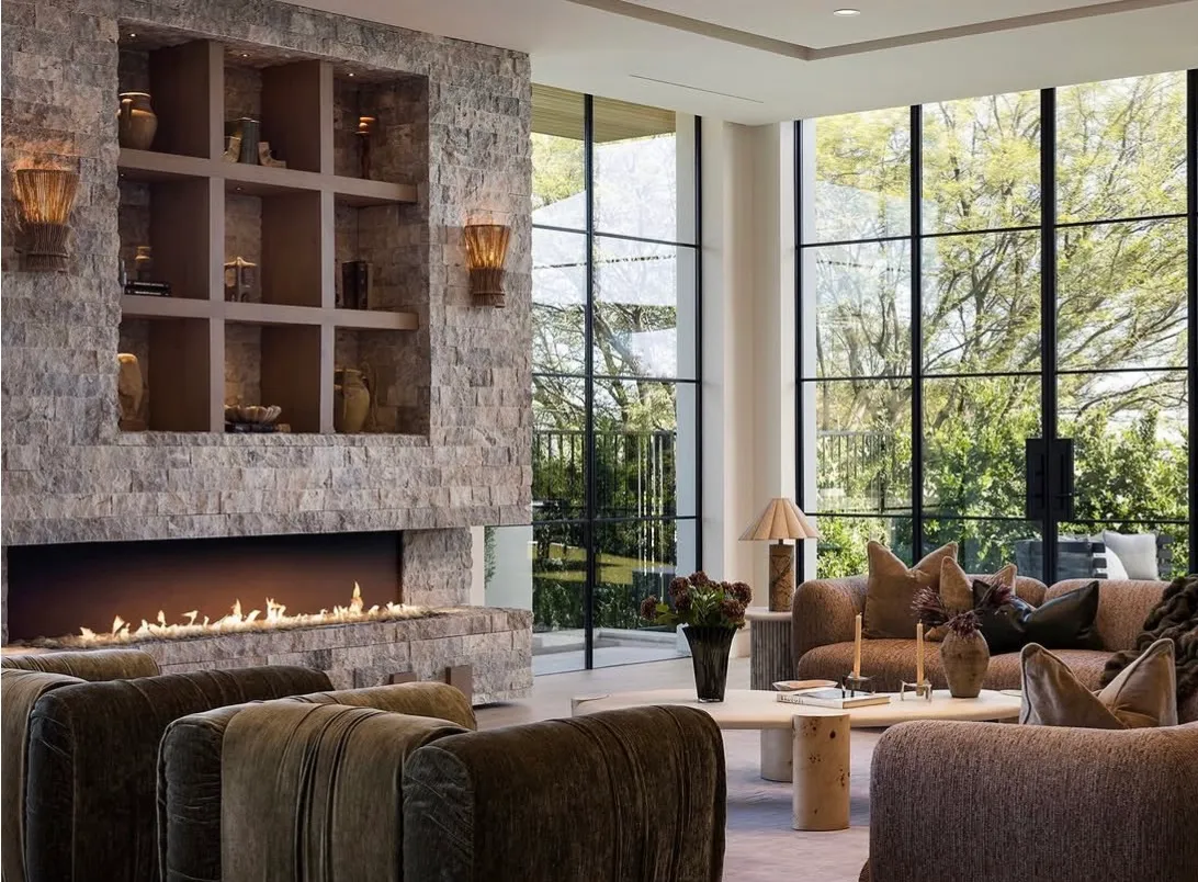 Double-height living room at The Opal in Beverly Hills — a full-height split-face stacked silver travertine fireplace with a built-in grid of recessed niche shelving and a long linear fire, flanked by floor-to-ceiling steel-and-glass windows looking onto mature trees, with velvet and bouclé seating below.
