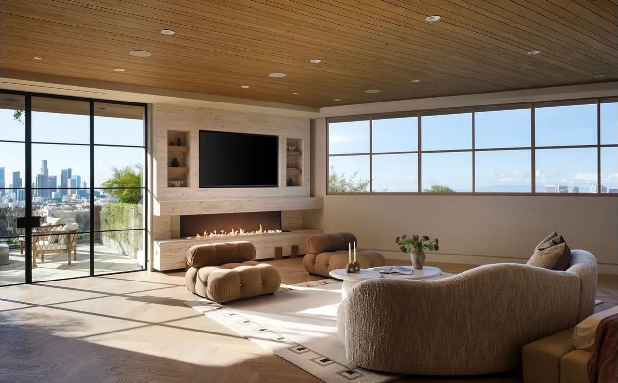 Living room at The Opal in Beverly Hills — a long horizontal travertine media wall in a soft ivory vein-cut field framing a linear gas fireplace and a recessed television, with paired niche shelves, a slatted wood ceiling, steel-framed glass walls opening to a terrace, and the downtown Los Angeles skyline beyond.