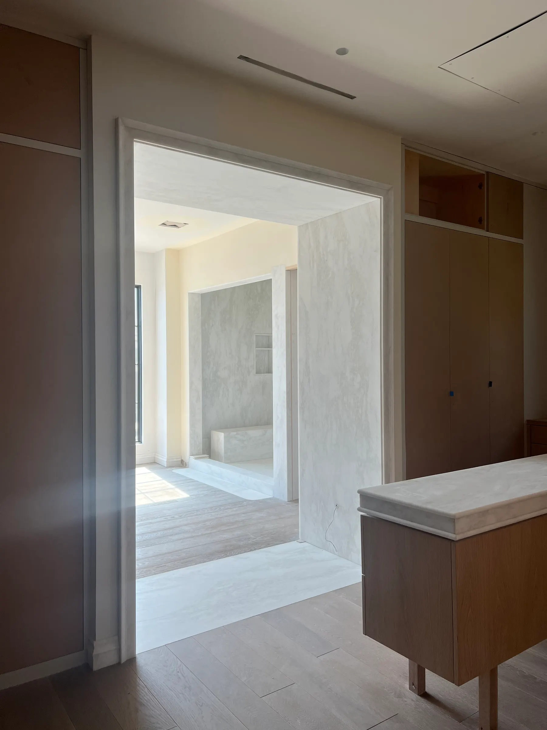 Primary bath suite viewed through a doorway clad in honed Rhino White marble — full-height slab jambs and head, with a marble-lined shower bench and threshold beyond, finished oak millwork and pale wide-plank flooring.