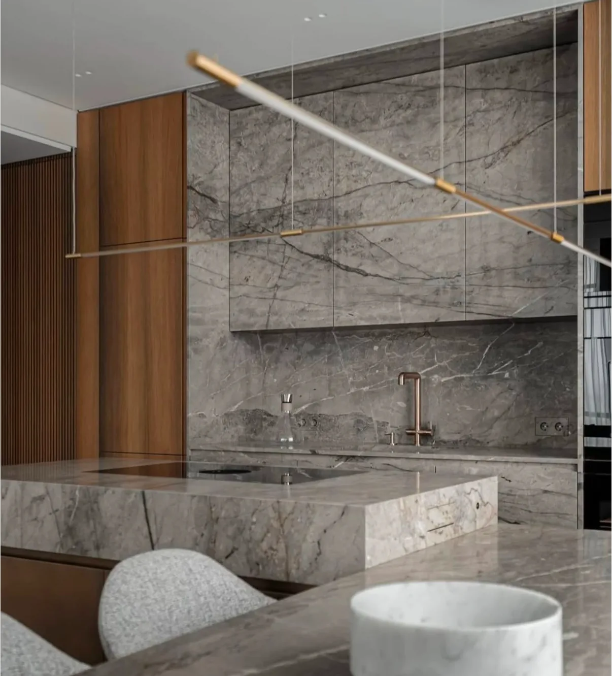 Minimalist kitchen with a full-height bookmatched Silver Roots taupe marble back wall and floating shelf in soft grey with charcoal veining, a monolithic taupe marble waterfall island with integrated cooktop, walnut cabinetry, a brushed-brass faucet, and a slim linear pendant.