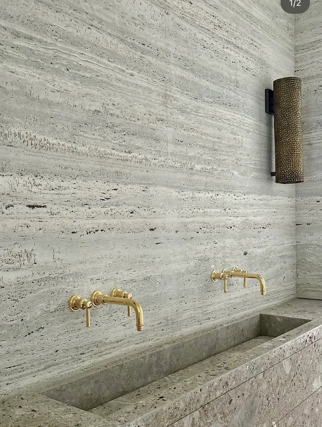 Vein-cut silver travertine bath wall with carved stone trough sink and unlacquered brass wall-mount faucets.