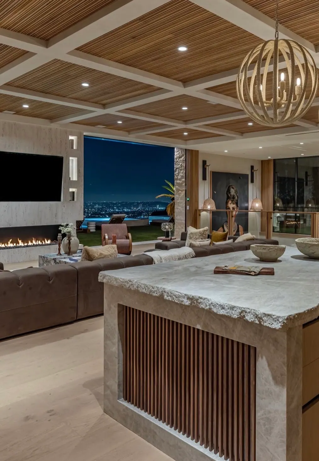 Great room with a long horizontal travertine fireplace surround and a live-edge natural travertine island, overlooking the Los Angeles skyline at night.