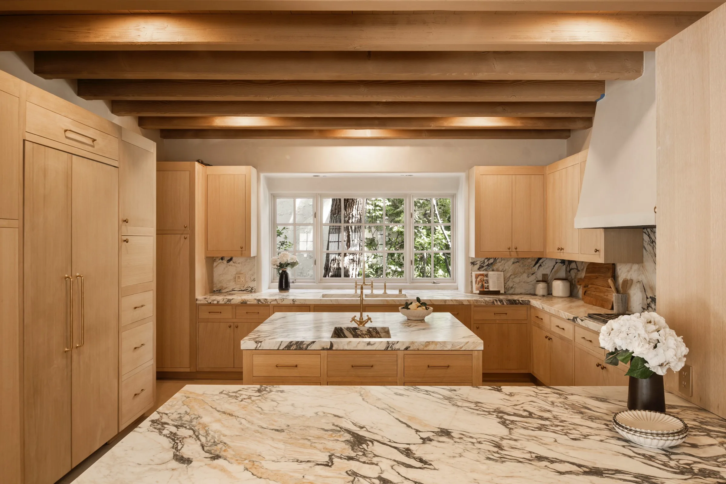 Warm white-oak kitchen with Antolini Calacatta Viola Extra marble throughout — perimeter counters and a full-height backsplash in an ivory field of plum, charcoal, and gold veining, a central island with an integrated marble bar sink and brass bridge faucet, a plaster hood, exposed Douglas-fir ceiling beams, and a casement window onto the trees.