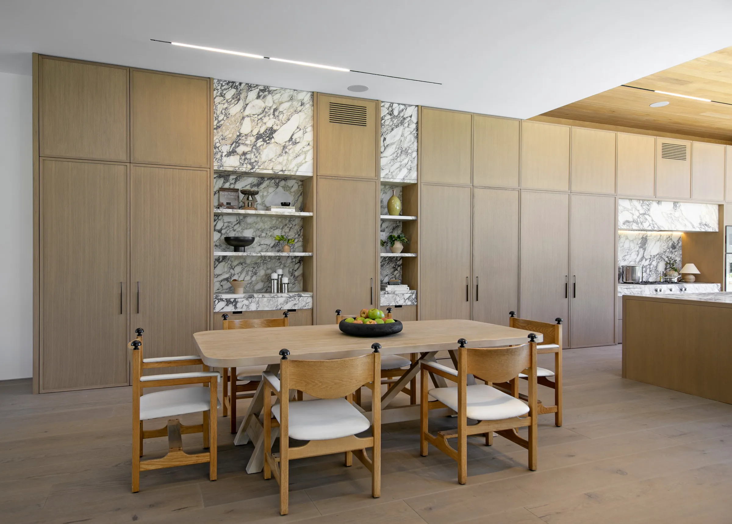 Dining area adjoining a Viola marble kitchen — two tall white-oak display niches lined in bookmatched Viola marble with floating marble shelves, a live-edge oak dining table with sculptural oak chairs, and the marble-backed kitchen island visible beyond.