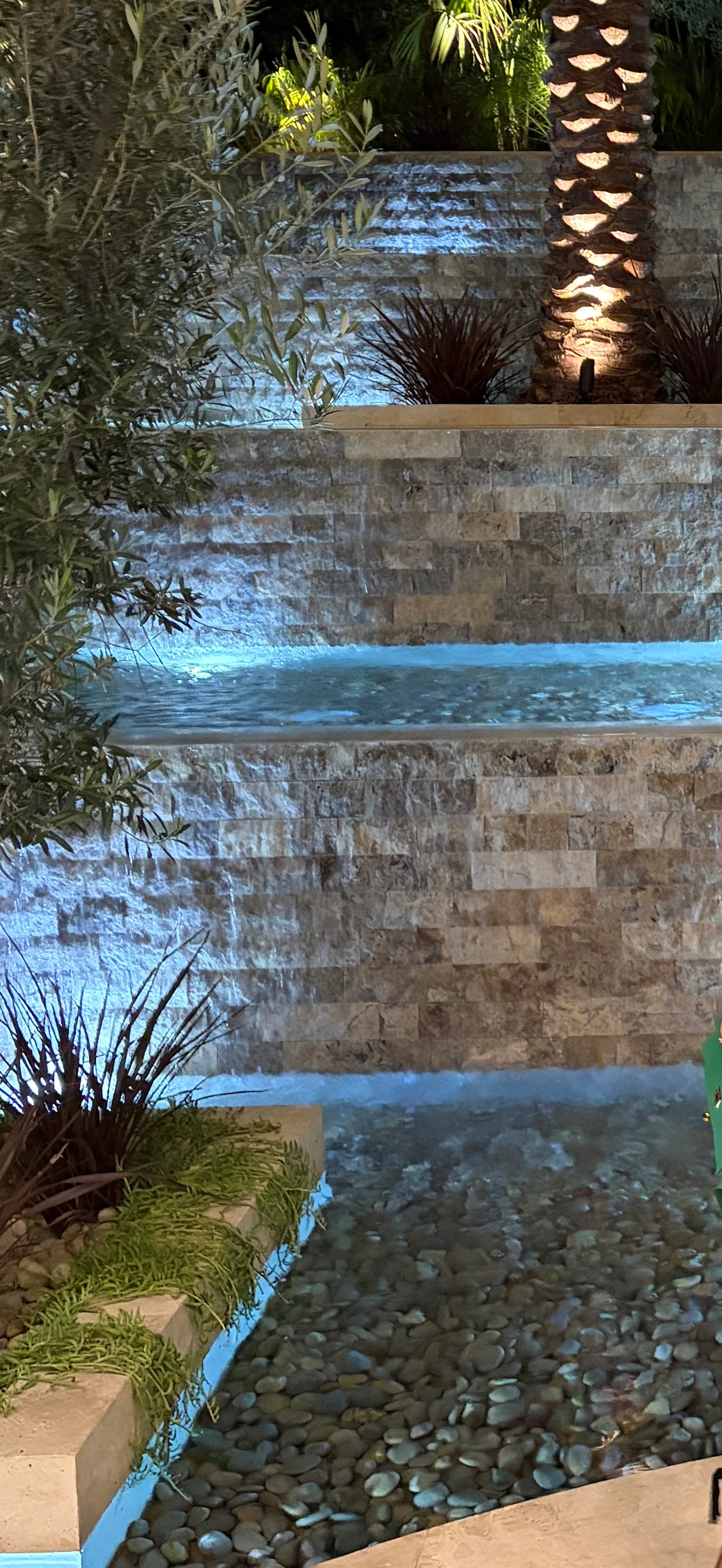 Silver Travertine split-face cladding rising behind a lit spillway and pool, with landscape palms in foreground