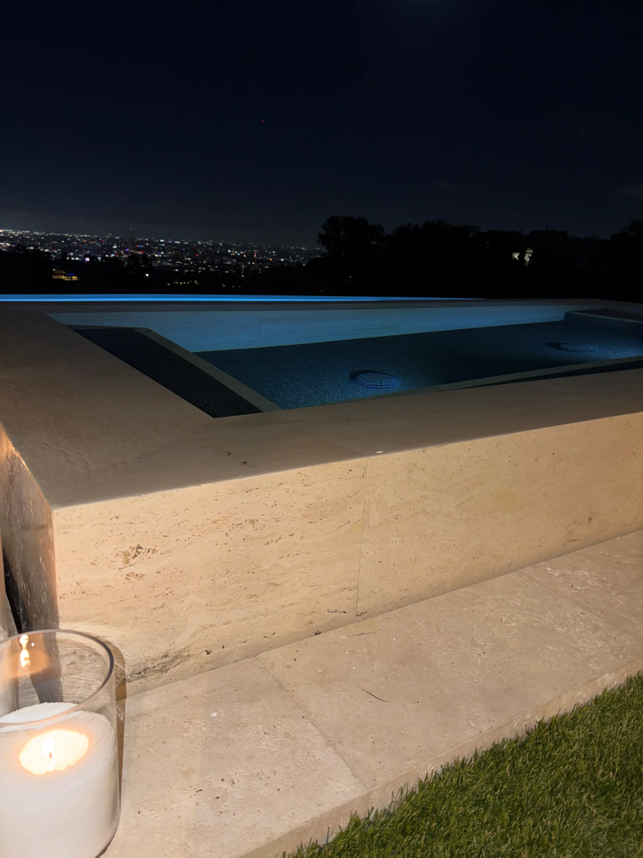 Unfilled travertine tile illuminated along a lawn edge at dusk, with candle and pool in background.